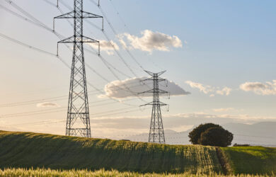 Tall electricity pylons with power lines stretch across a green field under a blue sky with scattered clouds; trees and distant mountains are visible in the background.