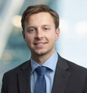 A young man in a suit and blue tie smiles at the camera in a professional setting, with a blurred background featuring soft lighting and modern architecture.