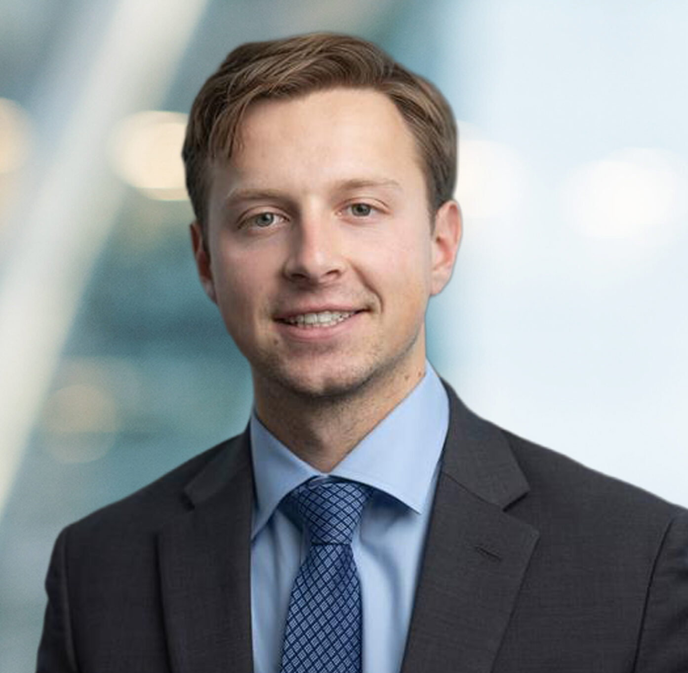 A young man in a suit and blue tie smiles at the camera in a professional setting, with a blurred background featuring soft lighting and modern architecture.