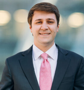 A man wearing a dark suit, white dress shirt, and pink tie is smiling at the camera. The background is blurred with lights and abstract shapes.