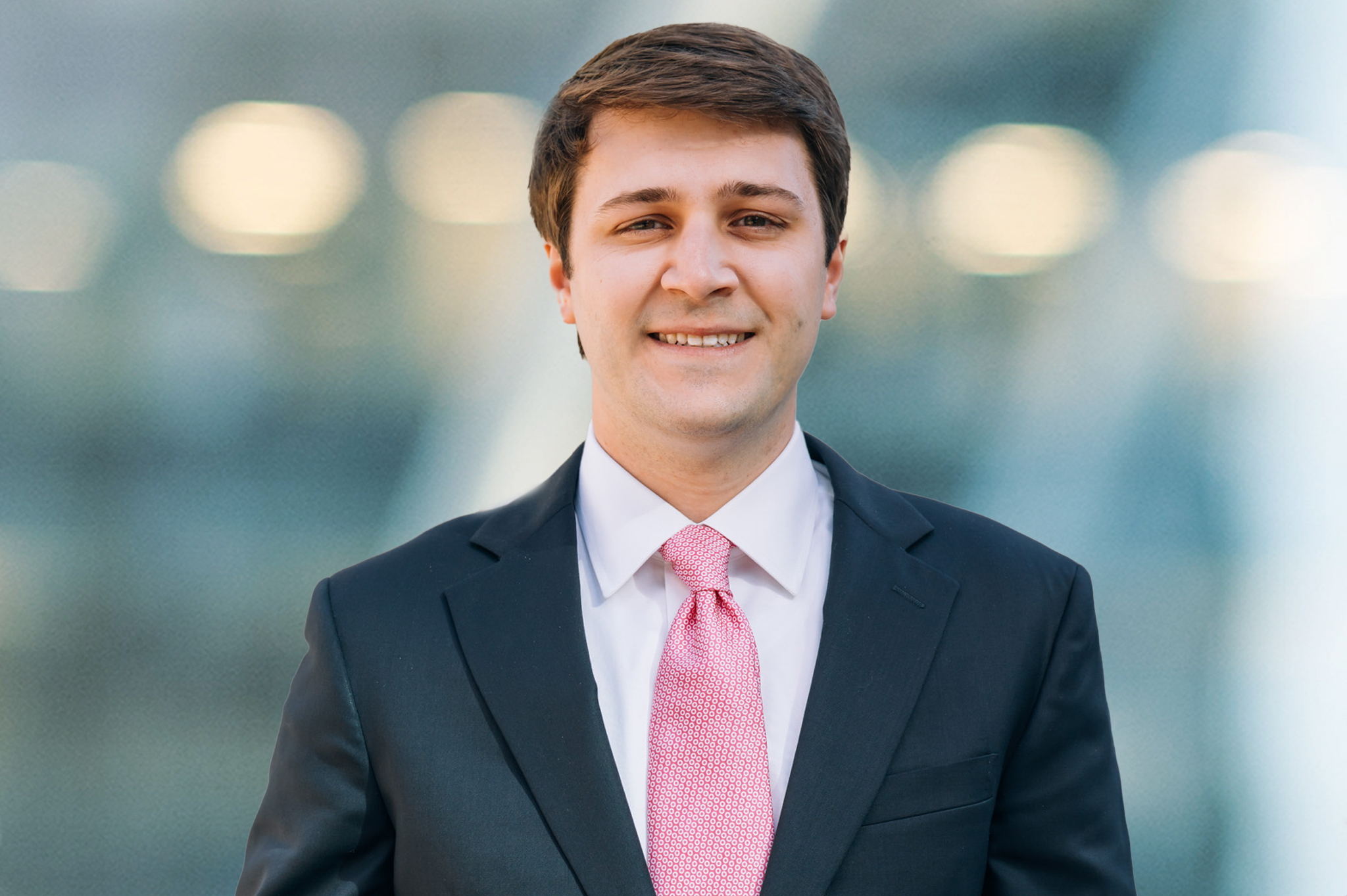 A man wearing a dark suit, white dress shirt, and pink tie is smiling at the camera. The background is blurred with lights and abstract shapes.