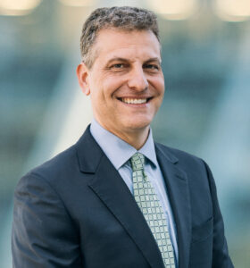 A man in a dark suit, light blue shirt, and patterned green tie smiles while standing outdoors in front of a blurred background.