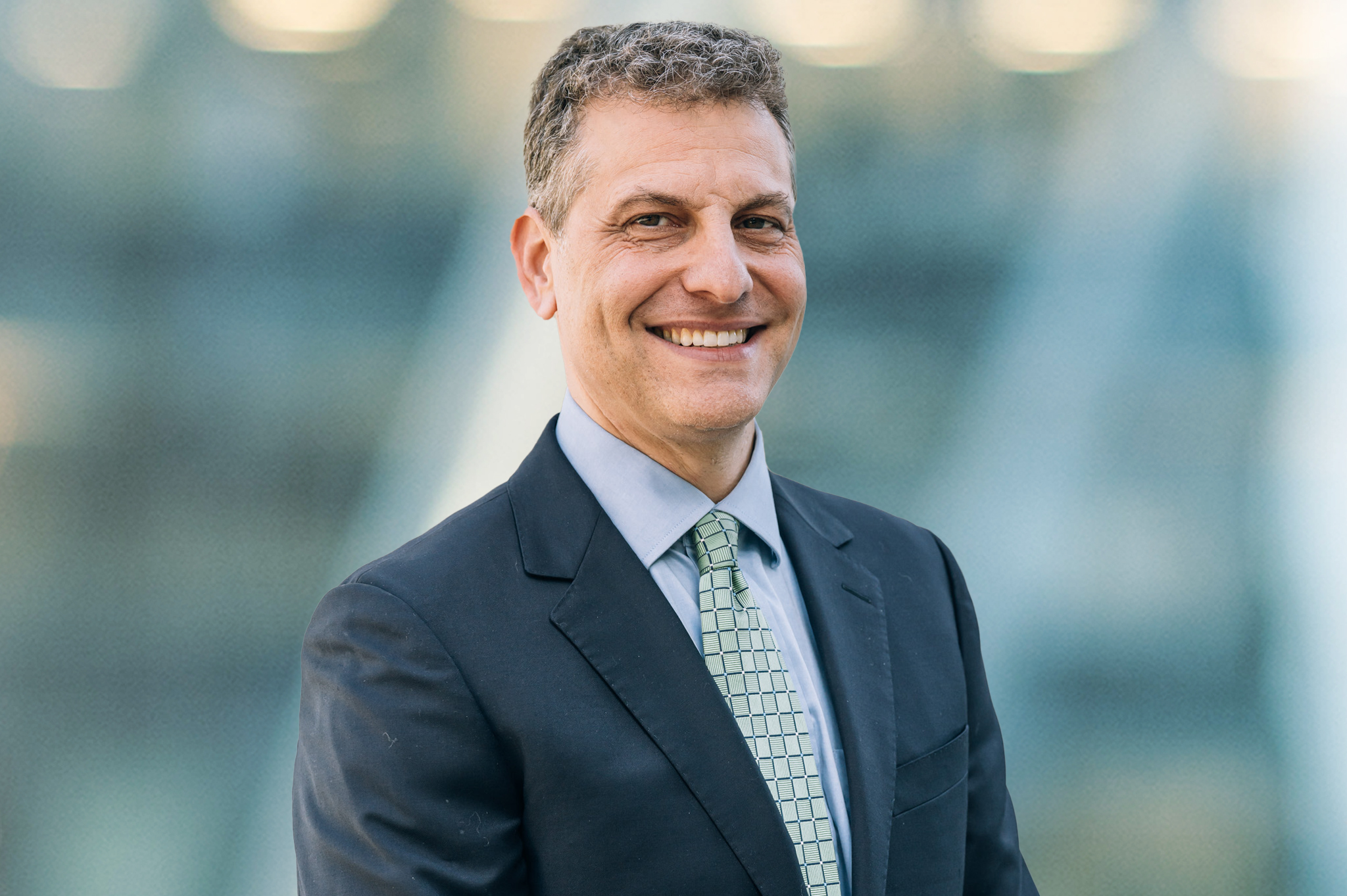 A man in a dark suit, light blue shirt, and patterned green tie smiles while standing outdoors in front of a blurred background.
