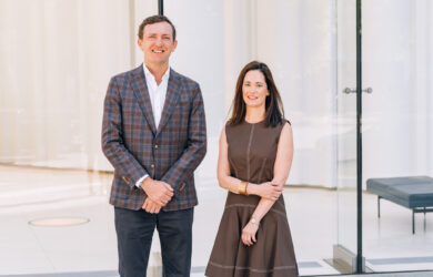 A man in a plaid blazer and dark pants stands next to a woman in a brown dress; both are smiling in front of large glass windows and a modern, minimalistic interior.