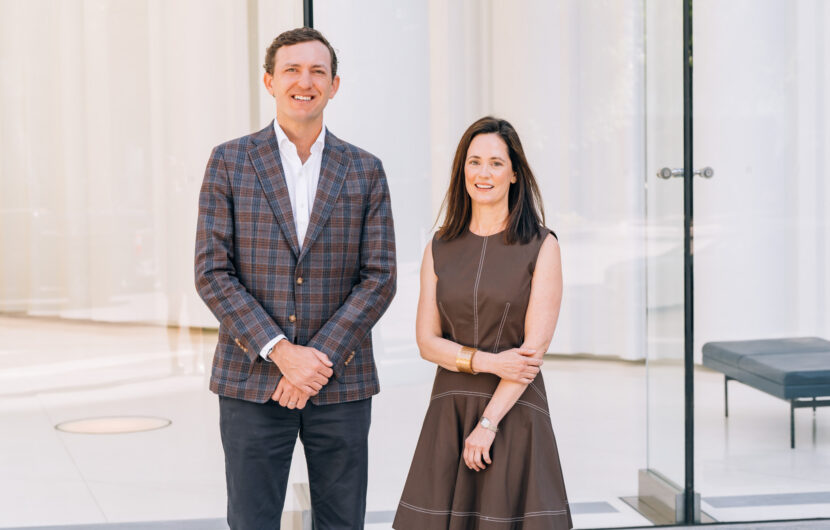 A man in a plaid blazer and dark pants stands next to a woman in a brown dress; both are smiling in front of large glass windows and a modern, minimalistic interior.