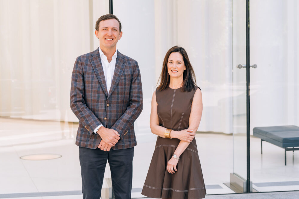 A man in a plaid blazer and dark pants stands next to a woman in a brown dress; both are smiling in front of large glass windows and a modern, minimalistic interior.