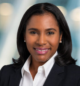 A woman with shoulder-length black hair, wearing a black blazer and white shirt, smiles at the camera against a blurred background with soft lights.