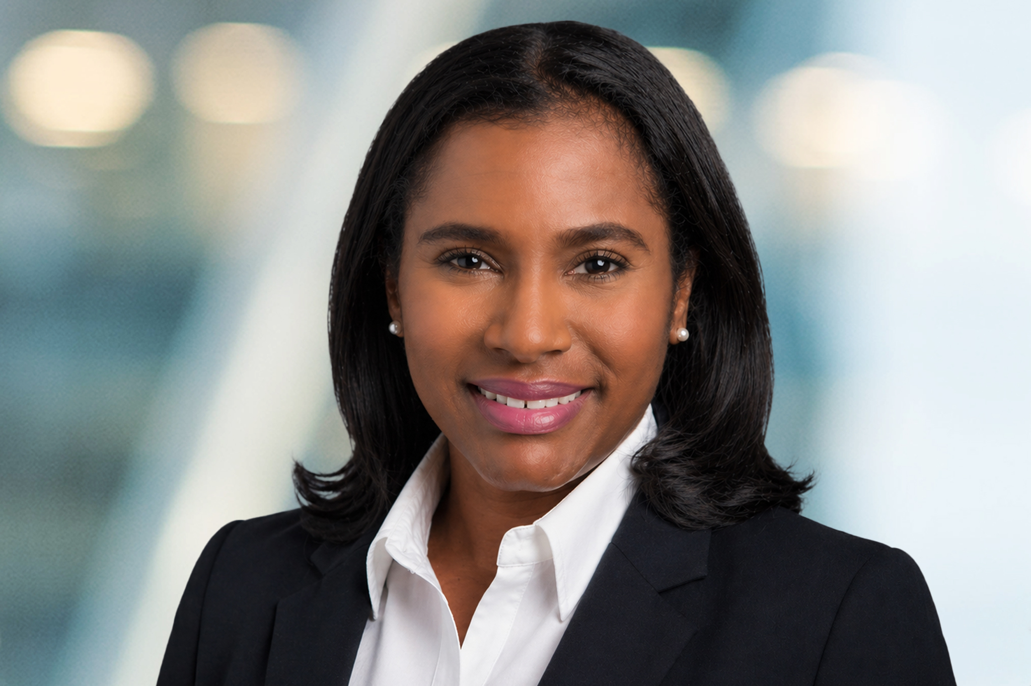 A woman with shoulder-length black hair, wearing a black blazer and white shirt, smiles at the camera against a blurred background with soft lights.