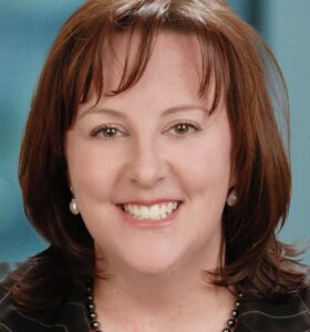 A woman with shoulder-length brown hair, wearing a black blazer with thin white stripes, a beaded necklace, and pearl earrings, smiling in front of a blue background.
