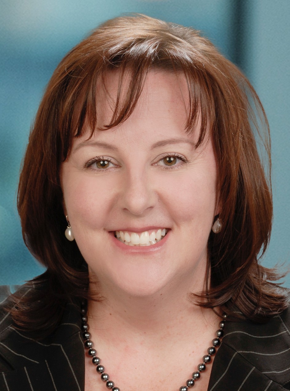 A woman with shoulder-length brown hair, wearing a black blazer with thin white stripes, a beaded necklace, and pearl earrings, smiling in front of a blue background.