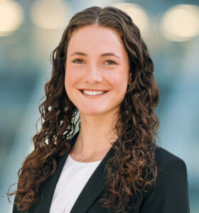 A young woman with long, curly brown hair smiles at the camera. She is wearing a black blazer over a white top, and stands in front of a blurred, light-blue background with circular lights.