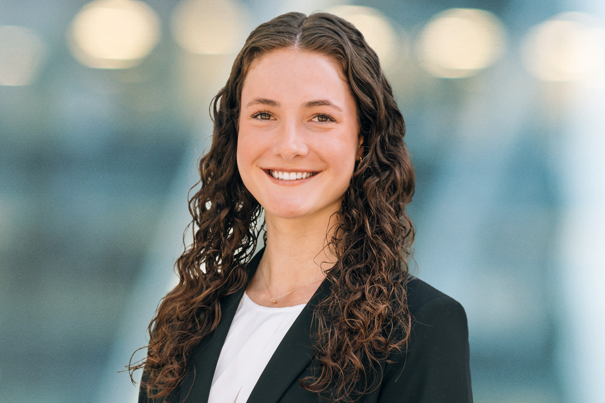 A young woman with long, curly brown hair smiles at the camera. She is wearing a black blazer over a white top, and stands in front of a blurred, light-blue background with circular lights.