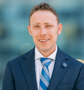 A man with short curly hair wearing a navy blue suit, white shirt, and blue striped tie stands in front of a blurred, modern glass background, smiling at the camera.