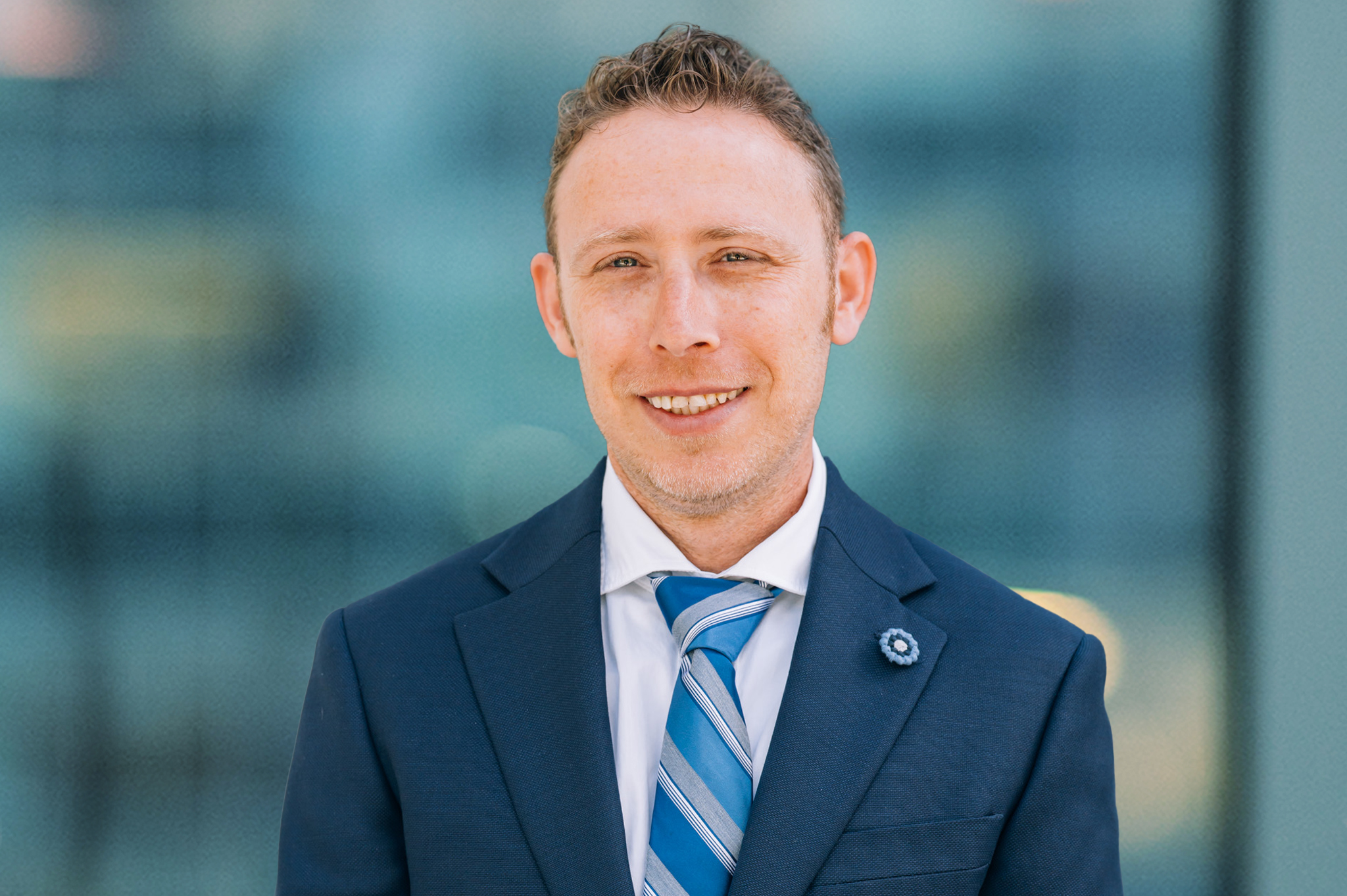 A man with short curly hair wearing a navy blue suit, white shirt, and blue striped tie stands in front of a blurred, modern glass background, smiling at the camera.