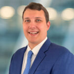 A man in a blue suit and tie smiles at the camera against a blurred blue and white background.