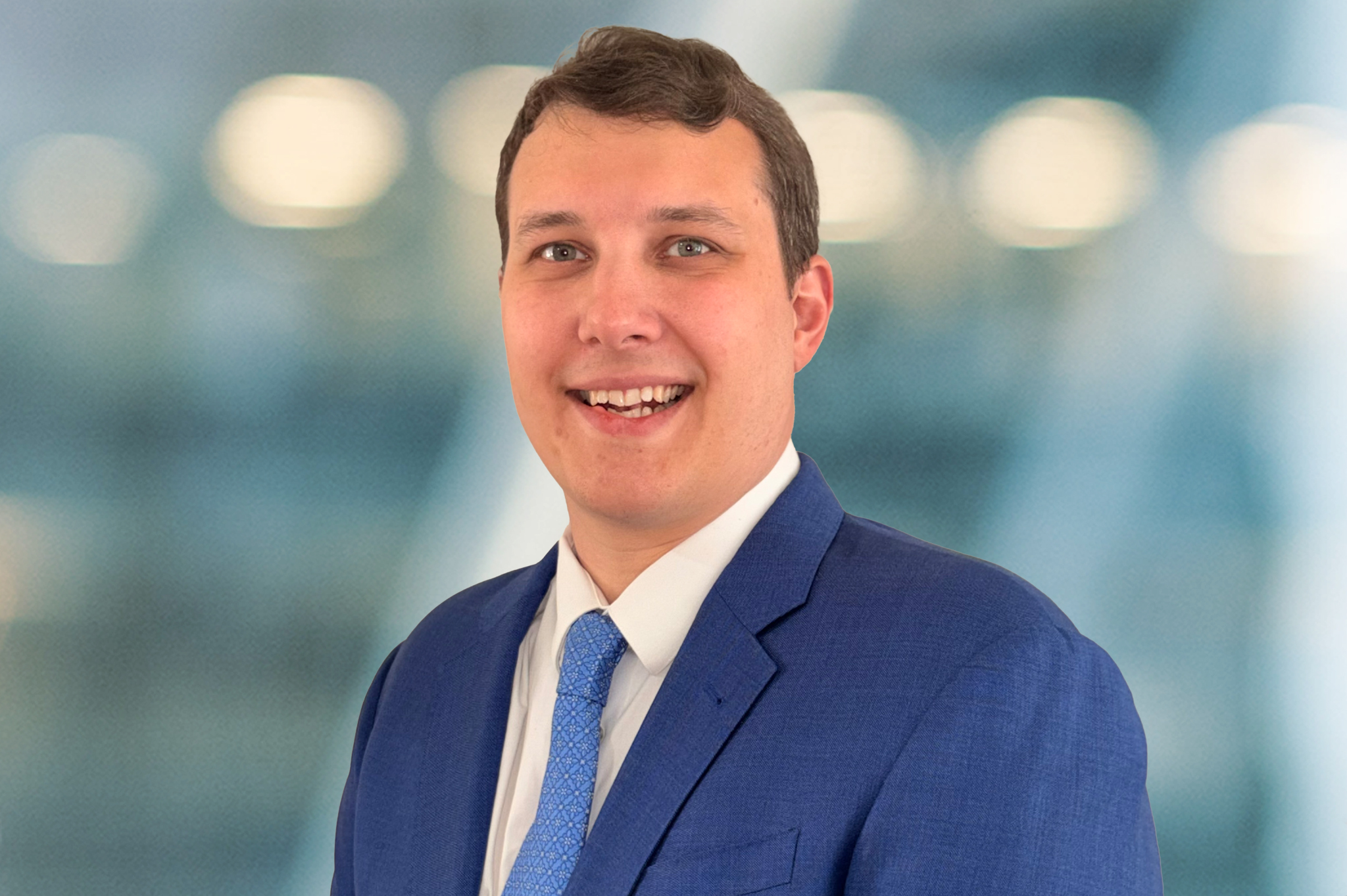 A smiling man in a blue suit, white shirt, and blue tie stands in front of a blurred, modern indoor background with circular lights.