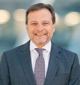 A middle-aged man with short brown hair and a trimmed beard, wearing a dark gray suit, white shirt, and patterned tie, smiling in front of a blurred blue-gray background.