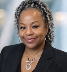 A woman with curly gray and black hair, wearing hoop earrings, a black blazer, and a distinctive silver necklace, smiles at the camera against a softly blurred background.