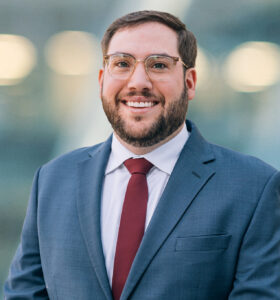 A man with short brown hair, a beard, and glasses smiles while wearing a blue suit, white shirt, and red tie. The background is blurred with soft lights.