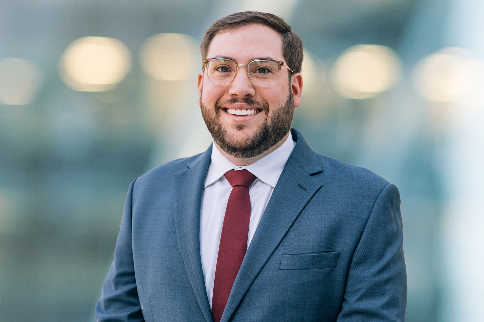A man with short brown hair, a beard, and glasses smiles while wearing a blue suit, white shirt, and red tie. The background is blurred with soft lights.