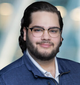 A man with glasses, dark hair, and a beard is smiling slightly. He is wearing a blue jacket over a white shirt. The background is blurred with cool tones and circular light spots.