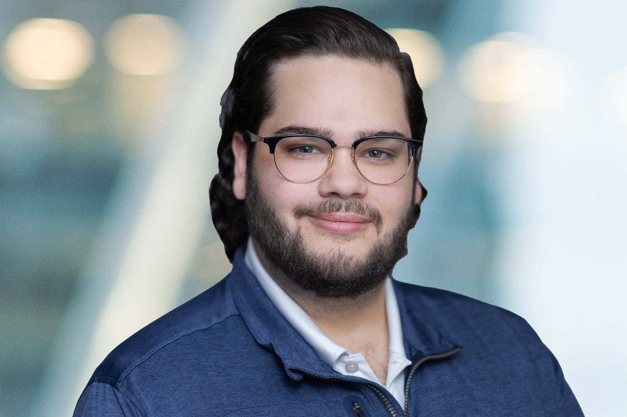 A man with glasses, dark hair, and a beard is smiling slightly. He is wearing a blue jacket over a white shirt. The background is blurred with cool tones and circular light spots.