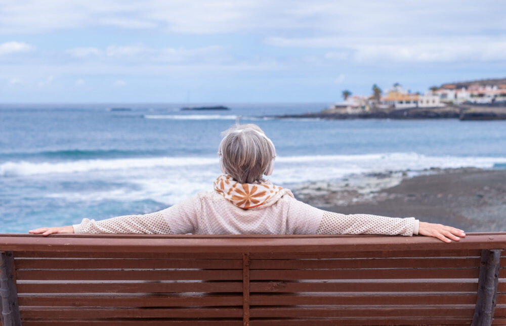 Rear view of senior woman wearing headphones sitting on outdoors bench at sea looking at horizon enjoying freedom and vacation. Active caucasian elderly woman relaxing on a sunny day close to the beach