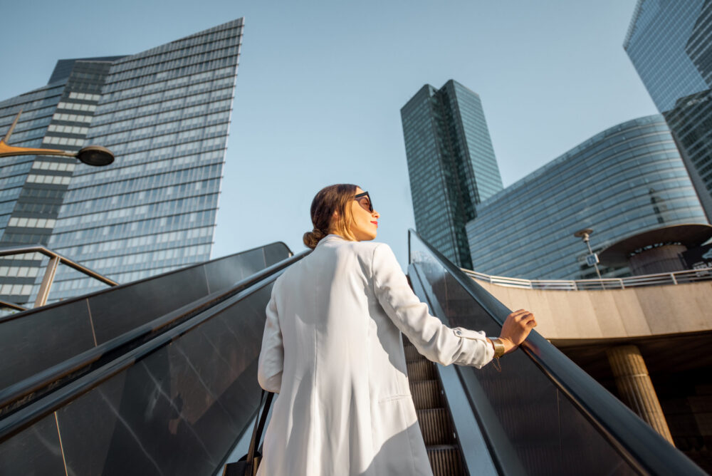 Stylish businesswoman in white suit going up on the escalator at the business centre outdoors with skyscrapers on the background in Paris