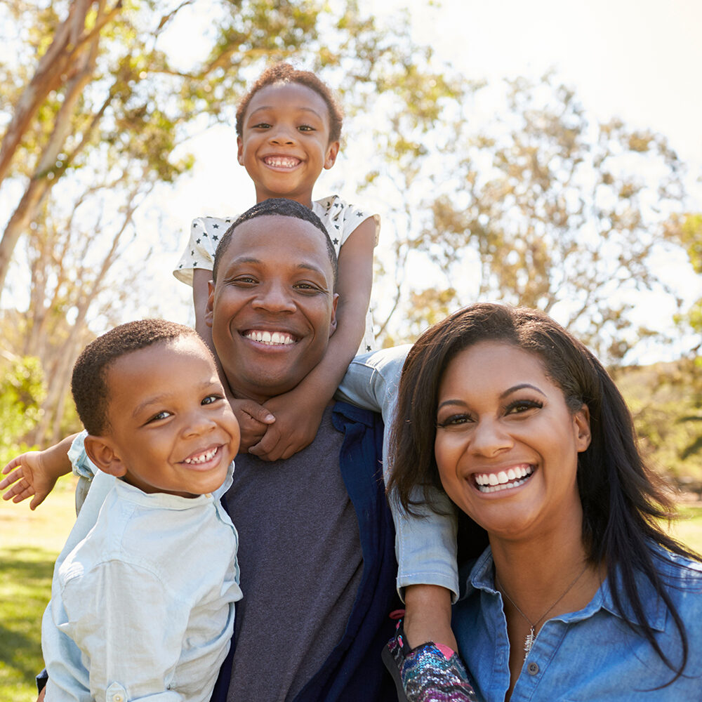 About-Us_put-the-client-first A smiling family of four poses outdoors on a sunny day. Two adults and two children are close together, with one child sitting on an adults shoulders. Trees and blue sky are visible in the background.