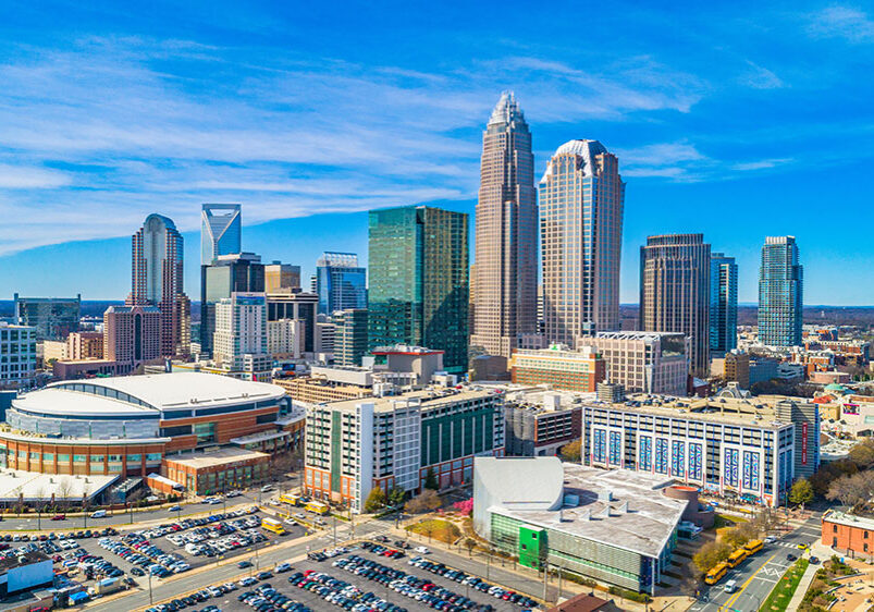 Aerial view of downtown Charlotte, North Carolina, featuring modern skyscrapers, the Spectrum Center arena, and surrounding buildings under a clear, blue sky.