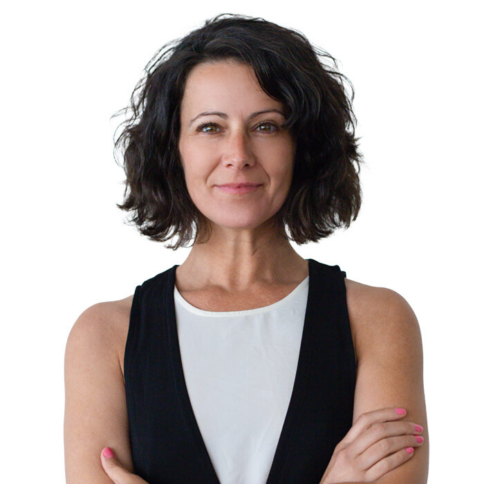 A woman with short, wavy dark hair stands with arms crossed and a confident expression. She is wearing a sleeveless black vest over a white top, and has pink-painted fingernails, against a plain white background.