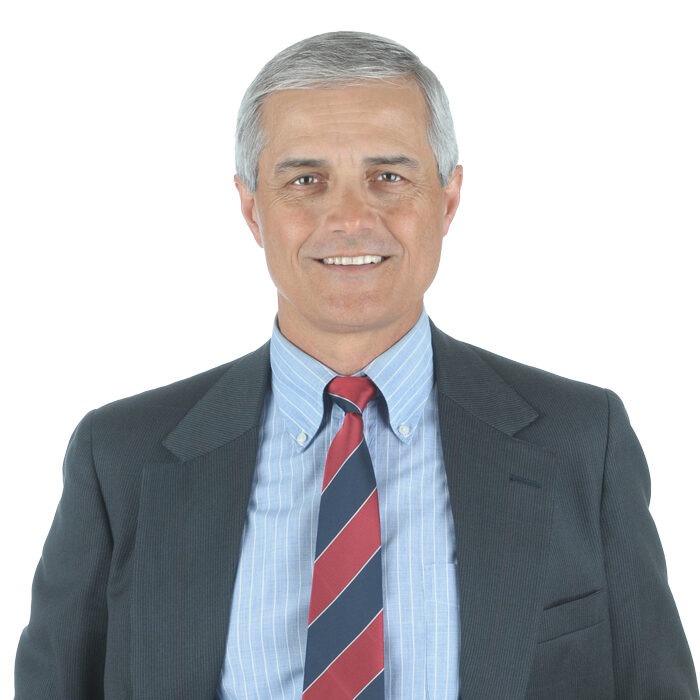 A middle-aged man with short gray hair, wearing a dark suit jacket, light blue shirt, and red and navy striped tie, smiling at the camera against a plain white background.
