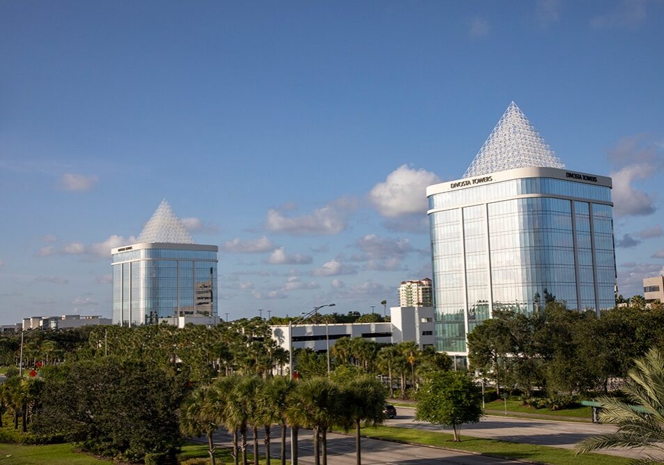 Two modern, glass office buildings with pyramid-shaped tops rise above palm trees and greenery under a blue sky with scattered clouds. The buildings are labeled Daytona Towers.