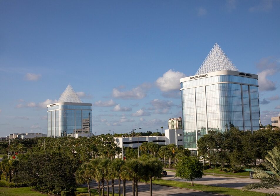 Two modern, glass office buildings with pyramid-shaped tops rise above palm trees and greenery under a blue sky with scattered clouds. The buildings are labeled Daytona Towers.