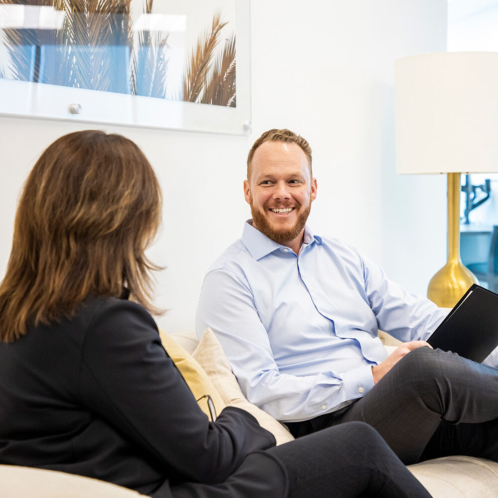 Our-Purpose_Do-the-Right-Thing A man and a woman sit on a sofa in a bright office, engaged in conversation. The man is smiling, holding a tablet, while the woman listens. A lamp and framed artwork are visible in the background.