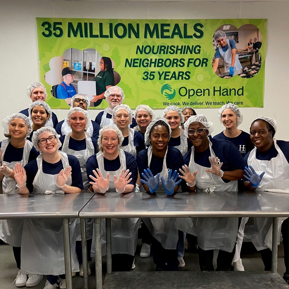Our-Purpose_Pour-Your-Heart-Into-It A group of smiling volunteers in hairnets, aprons, and gloves stand in front of a banner that reads “35 MILLION MEALS, NOURISHING NEIGHBORS FOR 35 YEARS, Open Hand.” They pose together in a kitchen setting.
