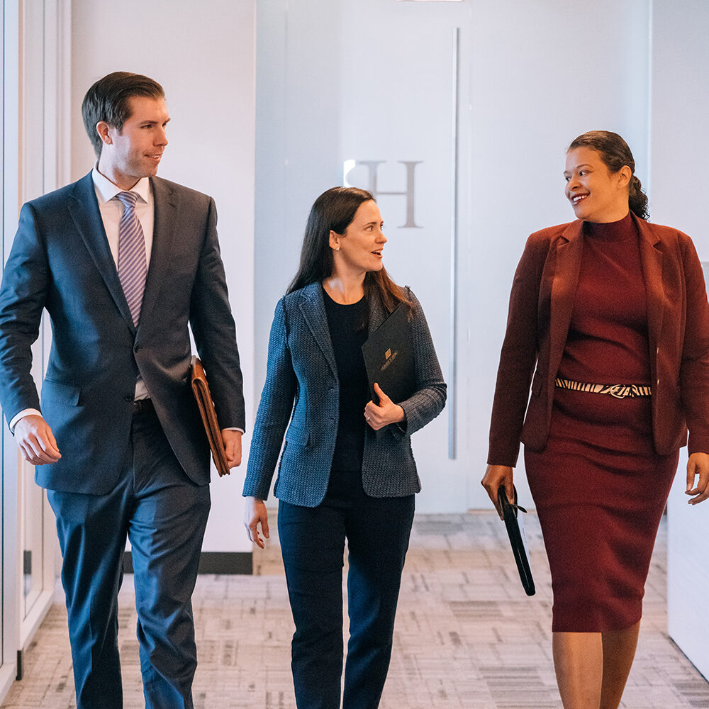 Our-Purpose_Raise-the-Bar Three professionally dressed people walk together down a hallway, smiling and holding folders, suggesting a business or office environment.