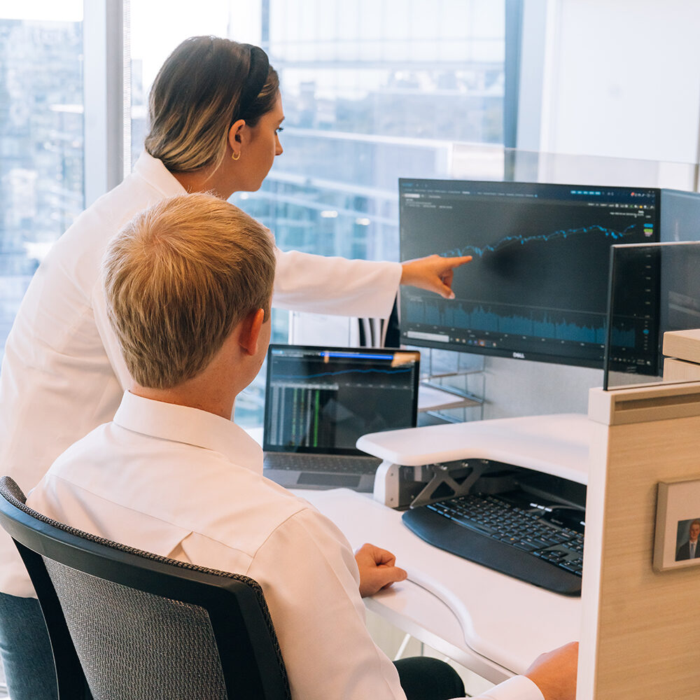 Our-Purpose_Work-As-A-Team Two people in business attire analyze a financial graph on a large computer monitor in a modern office, with one person seated and the other standing, pointing at the screen.