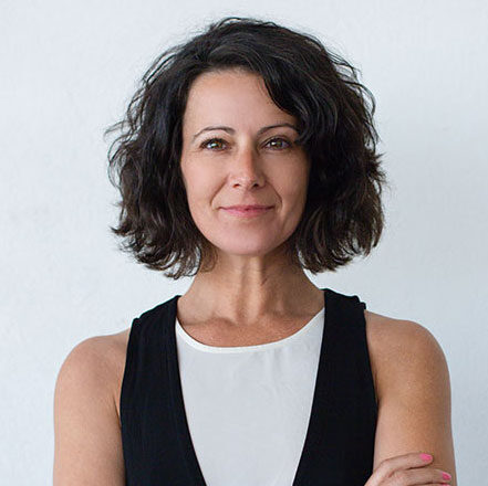 A woman with short, wavy dark hair stands confidently against a plain white background, wearing a sleeveless black vest over a white top and smiling slightly with her arms crossed.