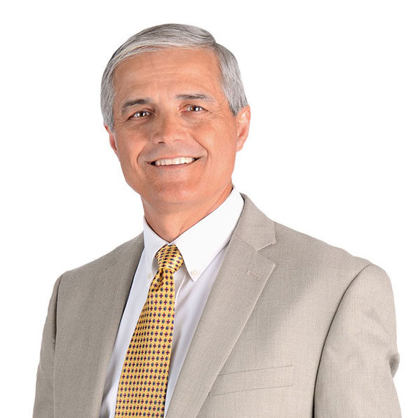 A smiling middle-aged man with short gray hair wearing a beige suit, white shirt, and yellow patterned tie, standing against a plain white background.