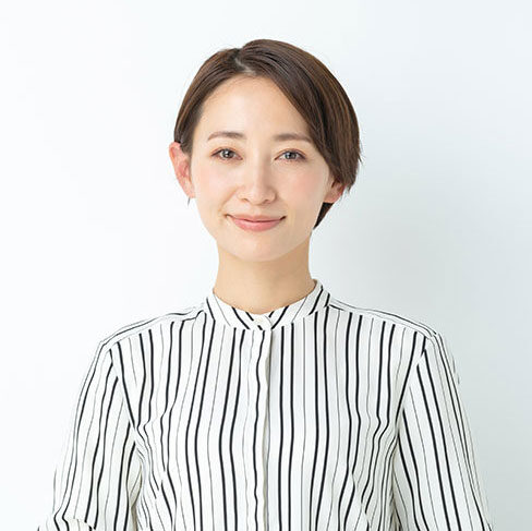 A woman with short brown hair smiles gently, wearing a white blouse with vertical black stripes, standing against a plain white background.