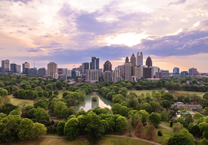 Aerial view of a city skyline at sunset, with tall buildings in the background and a large green park with trees and a small lake in the foreground. The sky is partly cloudy with soft sunlight.