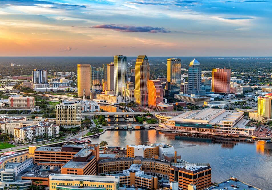 Aerial view of downtown Tampa, Florida at sunset, featuring modern high-rise buildings, waterfront structures, and the Hillsborough River reflecting colorful sky and city lights.
